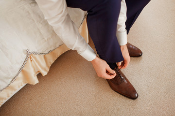 A man in a blue suit ties up shoelaces on brown leather shoes brogues on a wooden parquet background
