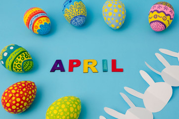 Colorful decorated eggs, white bunny paper chain and April made from colorful letters on blue background. Easter Day decoration