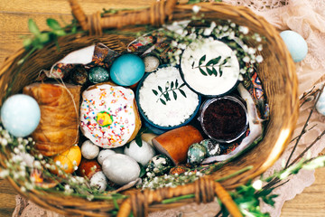 Stylish Easter eggs, easter bread cake, ham, beets, sausage, butter, green branches in wicker basket on rustic fabric with spring flowers. Happy Easter, holiday traditional feast. Top view