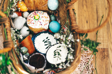 Stylish Easter eggs, easter bread cake, ham, beets, sausage, butter, green branches in wicker basket on rustic fabric with spring flowers. Happy Easter, holiday traditional feast. Top view