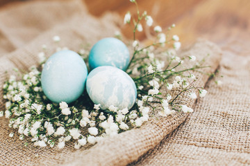 Stylish Easter eggs with spring flowers in floral nest on rustic fabric in sunny light on wood. Modern colorful eggs painted with natural dye in blue marble. Happy Easter, greeting card