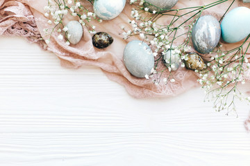 Stylish Easter eggs on rustic fabric with spring flowers and eucalyptus branch on white wooden background, flat lay. Space for text. Modern pastel eggs painted with natural dye. Happy Easter