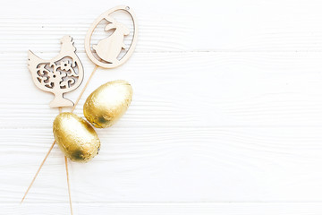 Fototapeta na wymiar Stylish Easter chocolate eggs in golden foil and simple wooden bunny and chicken decorations on white wooden background, flat lay with space for text. Happy Easter. Holiday gift