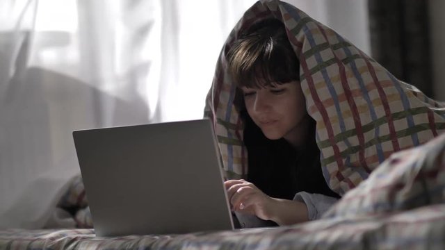 young woman lying on a bed covered with a blanket and typing on a laptop