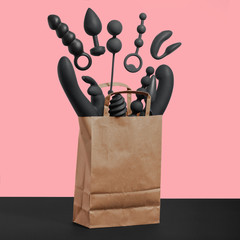 A paper shopping bag filled with black silicone sex toys on a pink background. Erotic toy for fun. Sex gadget and masturbation device. View from above. Place for text. Flat lay. Sex shop concept.