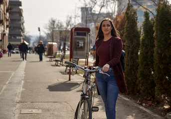 one young woman, 20-29 years old, pushing her old retro bicycle on a street in a city. pedestrians in distance (out of focus, blurred)