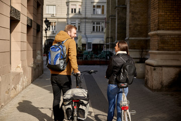 rear view, two friends talking and walking in a street, pushing their bicycles on a street in a city.