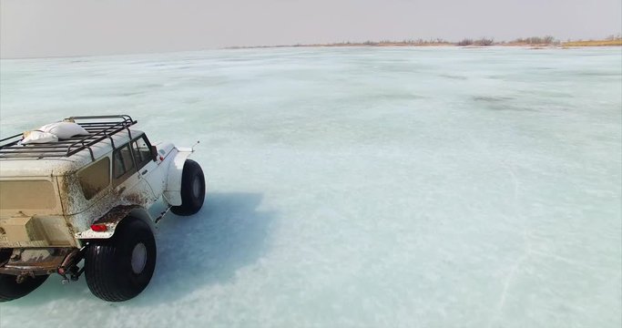 Dynamic aerial view of white swamp buggy driving on picturesque blue ice of huge frozen lake Hanka in Russia