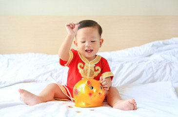 Happy little Asian baby boy in traditional Chinese dress putting some coins into a piggy bank sitting on bed at home. Kid saving money concept. Focus at piggybank.