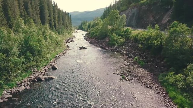 Close and slower forward drone shot over a river next to a road in a forest. Mountains are shown in the bachground. 4k quality video in Norway
