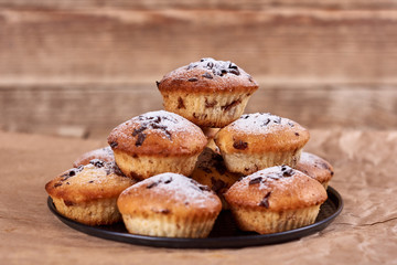 Cupcakes with chocolate chips with powdered sugar lie in a black baking sheet, on a kraft paper on a wooden background.