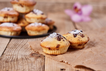 Obraz na płótnie Canvas The two cupcakes with chocolate chips with powdered sugar lie on a kraft paper next to other cupcakes in a black baking sheet and orchid flower on a wooden background.