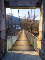 Obraz premium Concrete arch of a suspension bridge over the river. The early spring, the blue sky and mountains contrasts with yellowish vegetation and a wooden flooring of the bridge.
