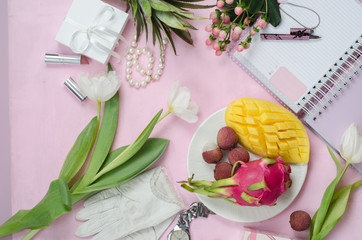 Women's accessories in white and pink colors whith tropical fruits. Mango, pineapple, pitahaya, lychee flowers, notebook with pen, cosmetics and gift boxes. Beauty women flat lay. Top view.