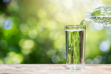 Pouring drinking water from bottle into glass on wooden tabletop on blurred fresh green nature background