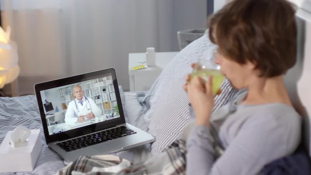 Lockdown of sick woman lying in bed, looking at computer screen, drinking hot drink from glass cup and communicating with male doctor by internet