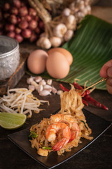 Thai fried noodles or pad thai with shrimp on black plate placed on the wood table there are eggs, garlic, bean sprouts, shallot, silver water bowl, chilli and cutting board placed back side. 
