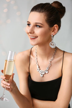 Young woman with beautiful jewelry and glass of champagne on light background