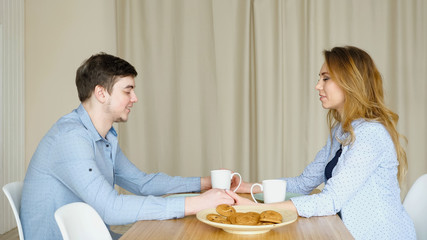blonde girl and guy sit at table across from each other hand in hand and pray before having breakfast against curtain