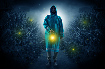 Man in raincoat at night coming from thicket and looking something with glowing lantern

