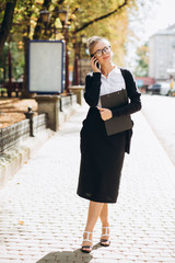 Beautiful trendy blond business woman in glasses with smartphone and folder of documents in her hands outdoors.