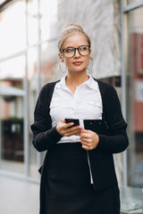 Beautiful trendy blond business woman in glasses talking by mobile phone and standing outside a office building.
