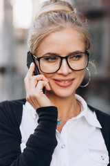 Beautiful trendy blond business woman in glasses talking by mobile phone and standing outside a office building.