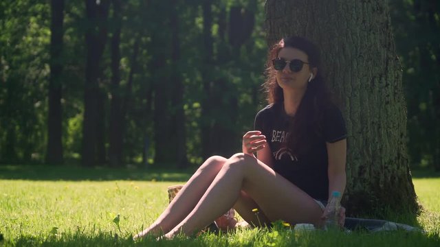 Young pretty woman with sunglasses uses smartphone and drinks mineral water from plastic bottle. Student girl sitting on grass in shade of tree in  park among  trees and foliage on bright sunny day.