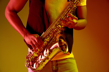 Obraz na płótnie Canvas African American handsome jazz musician playing the saxophone in the studio on a neon background. Music concept. Young joyful attractive guy improvising. Close-up retro portrait.