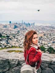 Brunette girl with long hair and a red jacket stands in the background of the panorama of San Francisco with twin Peaks hill