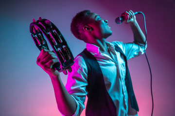 African American handsome jazz musician playing tambourine and singing into the microphone in the studio on a neon background. Music concept. Young joyful attractive guy improvising. Close-up retro - obrazy, fototapety, plakaty