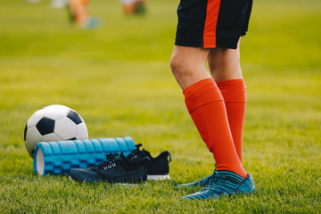 Legs of a football player boy in boots cleats with a ball and foam roller on the green lawn of the...