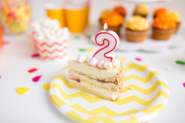 anniversary, celebration and festive concept - piece of cake with candle in shape of number two on plate at birthday party