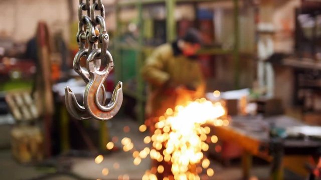 Construction plant. A big industrial lifting chain with a hook on the end hanging in the air. A man welding on the background