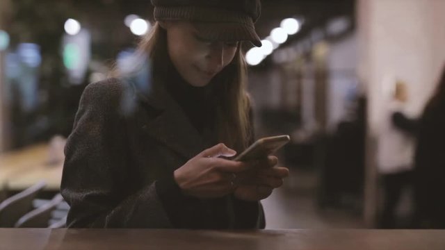 portrait of young beautiful fashionable brunette woman sitting in coffeehouse near window at evening and using phone. Model wearing stylish gray coat, hat and black golf. Seasonal Female fashion
