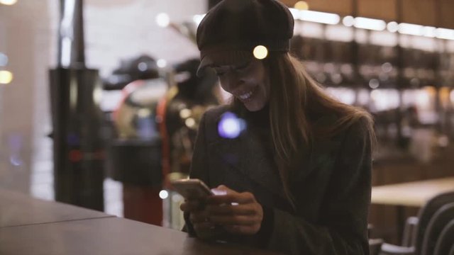 portrait of young beautiful fashionable brunette woman sitting in coffeehouse near window at evening and using phone. Model wearing stylish gray coat, hat and black golf. Seasonal Female fashion