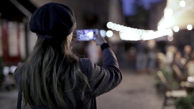 HD stock footage of young beautiful fashionable brunette woman standing on the street at night and taking photo or video using phone.