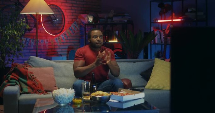 African American young man sitting on the couch with beer and pizza and worrying while watching American football game on TV in the dark room.
