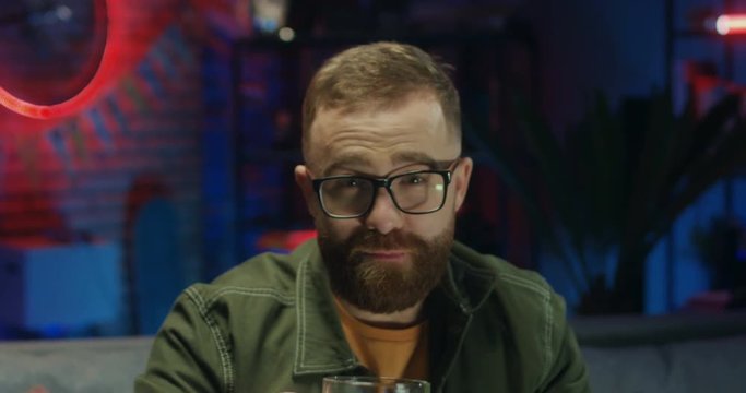 Portrait of the young cheerful Caucasian man in glasses drinking beer and doing cheers gesture to the camera and smiling in the dark room. Close up.