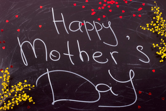 Happy Mother's Day written on a black chalkboard in white font next to the sprigs of yellow mimosa. Top view, flat lay.