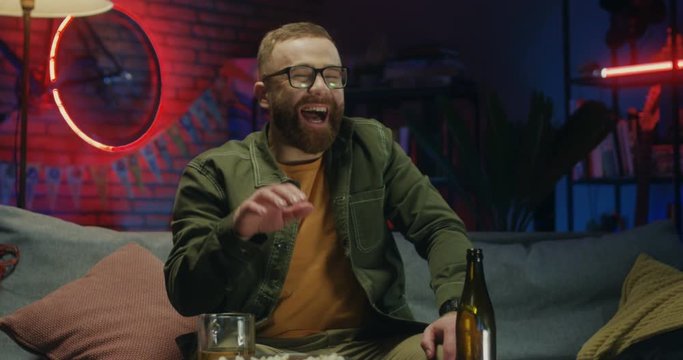 Caucasian red-haired young man in glasses sitting on the couch in the dark room at night, drinking beer and laughing while watching comedy movie on TV.