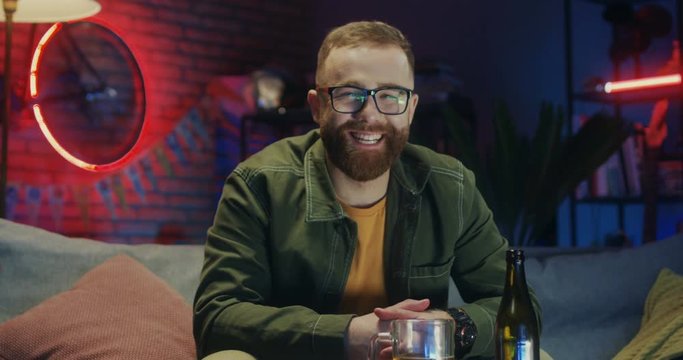 Portrait of the Caucasian joyful red-haired young man in glasses smiling to the camera and being in good mood while sitting on the sofa in the dark room.