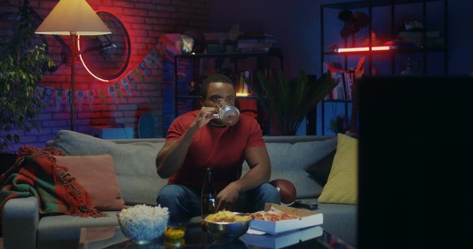 Young african American handsome man drinking beer and resting while watching sport channel on TV at home at night.