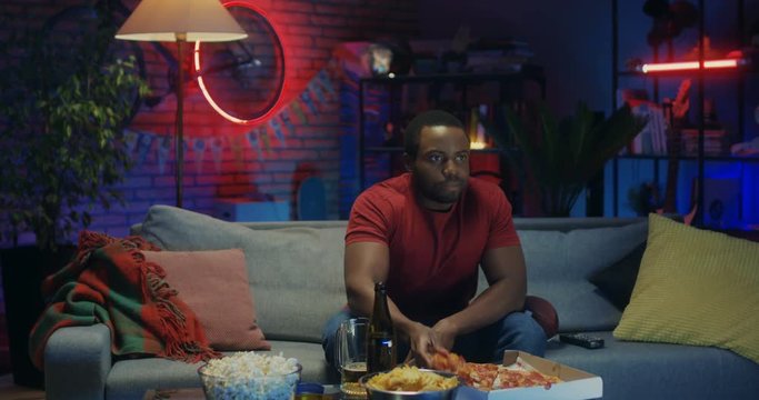 African American young attractive man sitting on the sofa in the living room at night, eating pizza and watching TV.