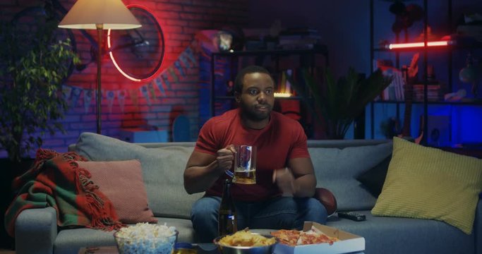 Good looking young African American guy drinking beer and cheering up for his favorite team while watching sport match on TV at night.