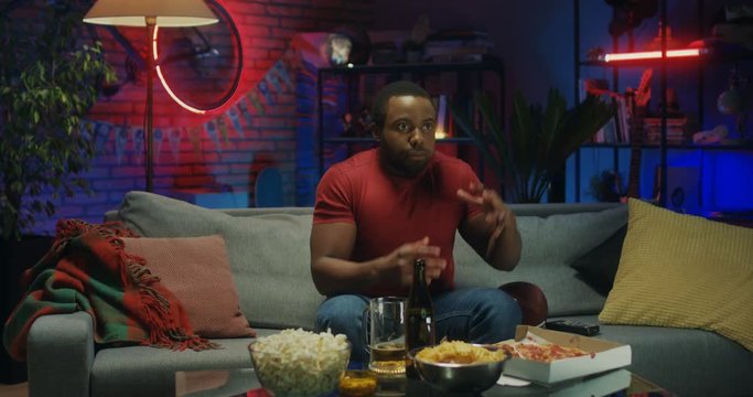 African American young man eating pizza and watching American football on TV in the dark room at night. then being upset as his team loosing.