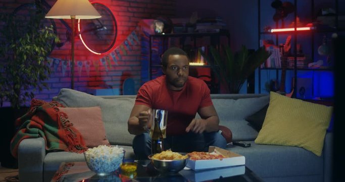 African American male sport fan watching football game on TV in the evening and drinking beer. At home.