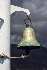 Bell hanging on sailing ship - Image. Ancient shiny brass bronze bell covered with patina on board the ferry with grey cloudy stormy sky on background . Sign of danger, storm.