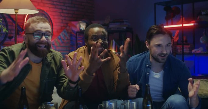 Close up of the joyful three multiethnic guys watching sport championship on TV at home in the evning and cheering up.