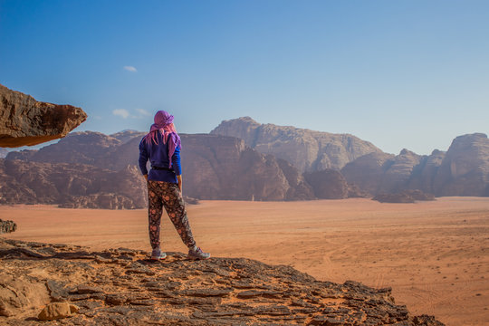 travel photography concept of girl in hijab stay back to camera on top of bare rock in Wadi Rum heritage touristic Jordanian place and looking on a beautiful dry scenery landscape with valley and rock